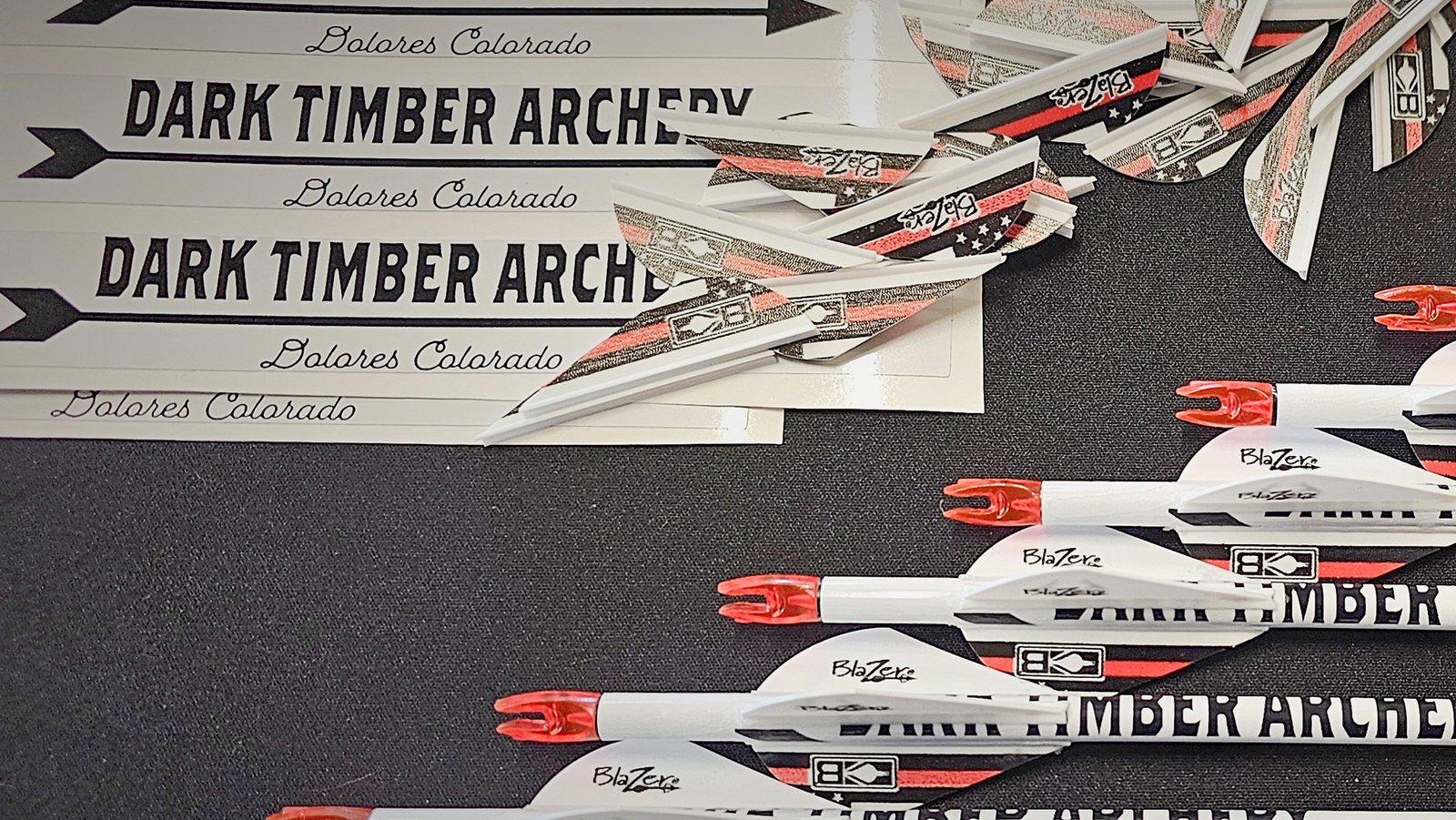 dark-timber-archery-arrow-services Custom arrows and Blazer vanes assembled by Dark Timber Archery in Dolores, Colorado, featuring branded wraps and arrow components on display.
